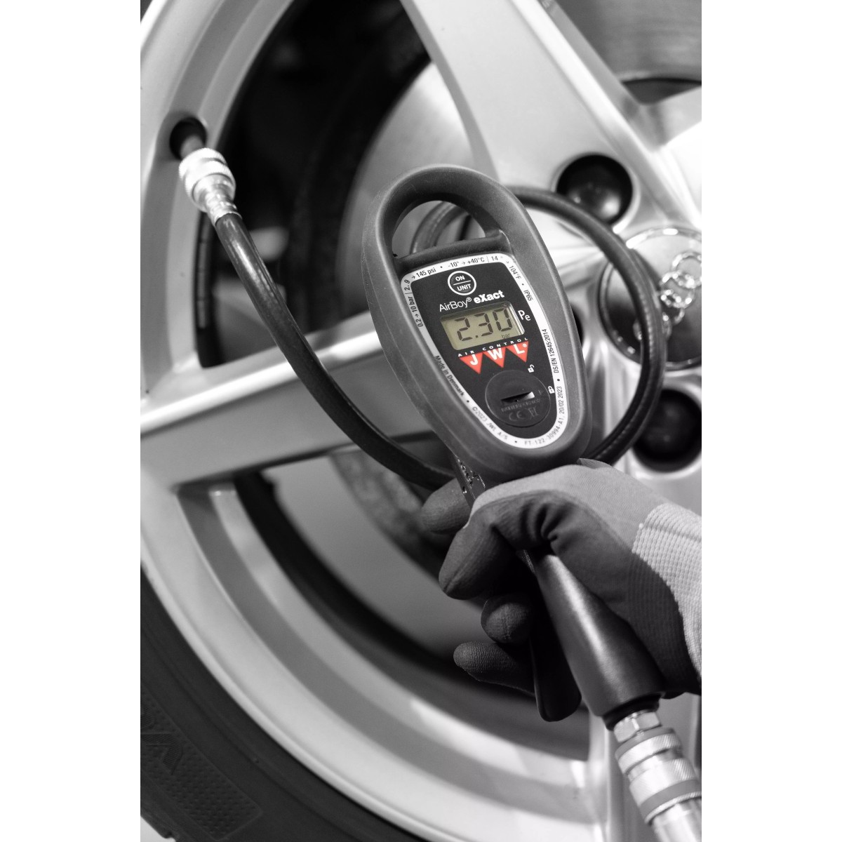 Reifenfüller JWL DIGITAL 0-10 bar Schwarzweißfoto: Hand im Handschuh hält digitales Reifenfüllgerät vor Autofelge; Schlauch ist am Ventil angeschlossen.