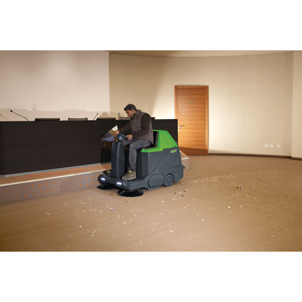 Aufsitz-Kehrsaugmaschine AUKM 600 Person fährt die grün-schwarze Aufsitz-Kehrmaschine in einem Konferenzraum.