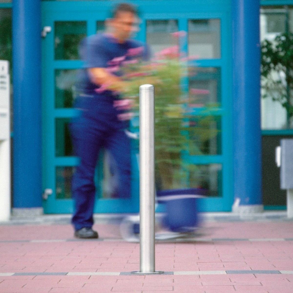 Edelstahlpfosten auf Pflaster; im Hintergrund unscharf eine Person vor blauem Eingang, neben einem rollenden Pflanzgefäß.