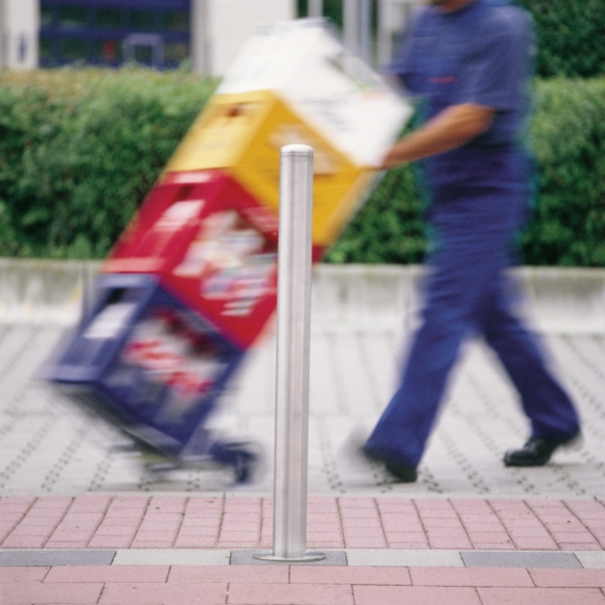 Edelstahlpfosten auf Pflaster; im Hintergrund unscharf eine Person, die eine Sackkarre mit Kartons schiebt.