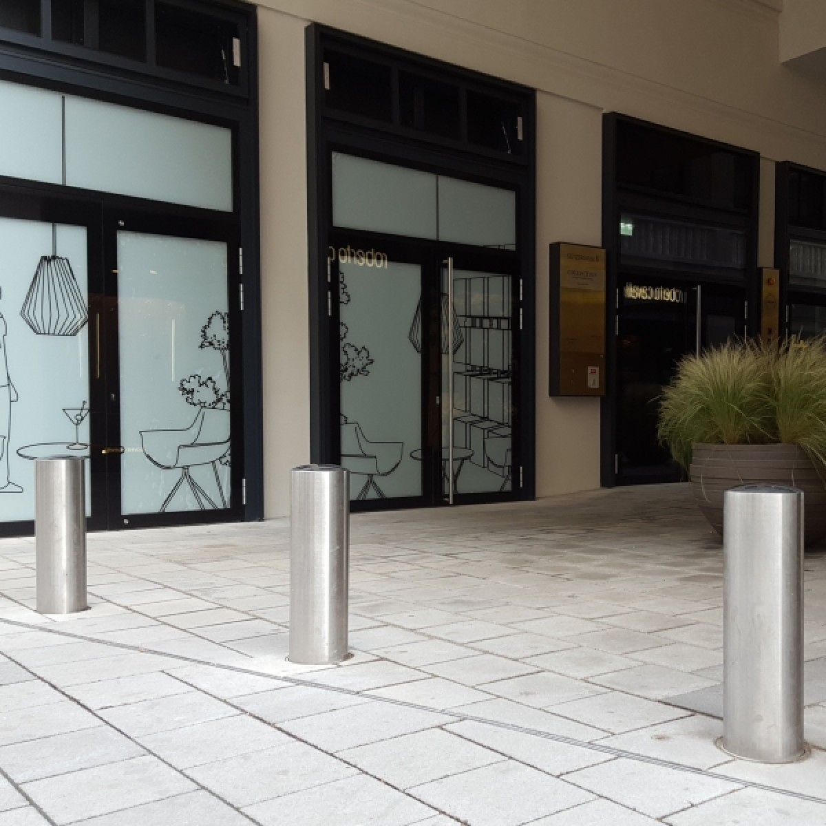Drei Edelstahlpoller vor Gebäudeeingang mit Glasfront; Pflasterfläche und großer Pflanzkübel rechts.