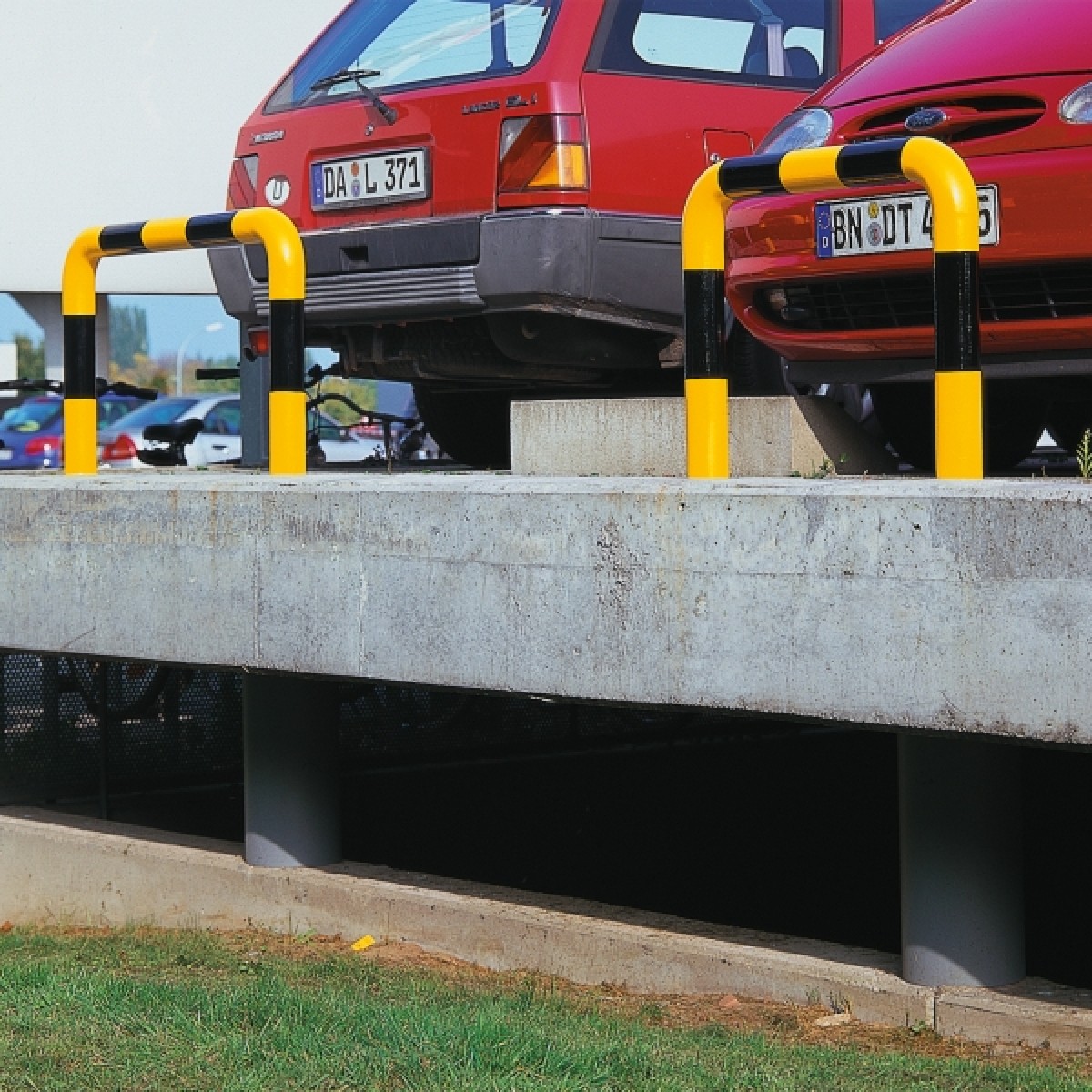 Rammschutz-Bügel - Ø 76 Foto: Zwei gelb-schwarz markierte Rammschutzbügel an einer Betonrampe vor parkenden roten Autos, Außenbereich.