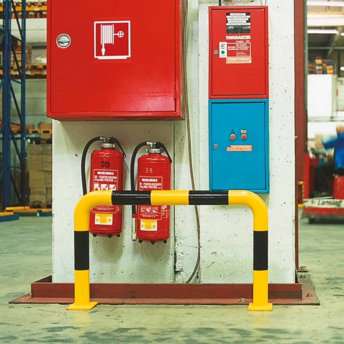 Rammschutz-Bügel - Ø 76 Gelb-schwarz gestreifter Rammschutzbügel vor Betonpfeiler; dahinter rote Wandkästen und zwei Feuerlöscher in Werkhalle.