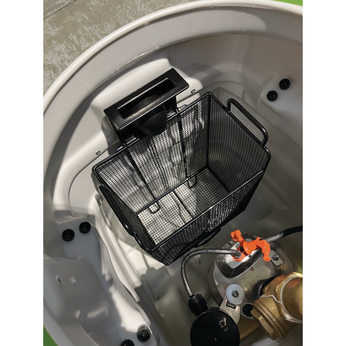 Innenansicht des Behälters mit Drahtkorb, Ansaugöffnung sowie sichtigen Schlauch- und Ventilanschlüssen am Boden.